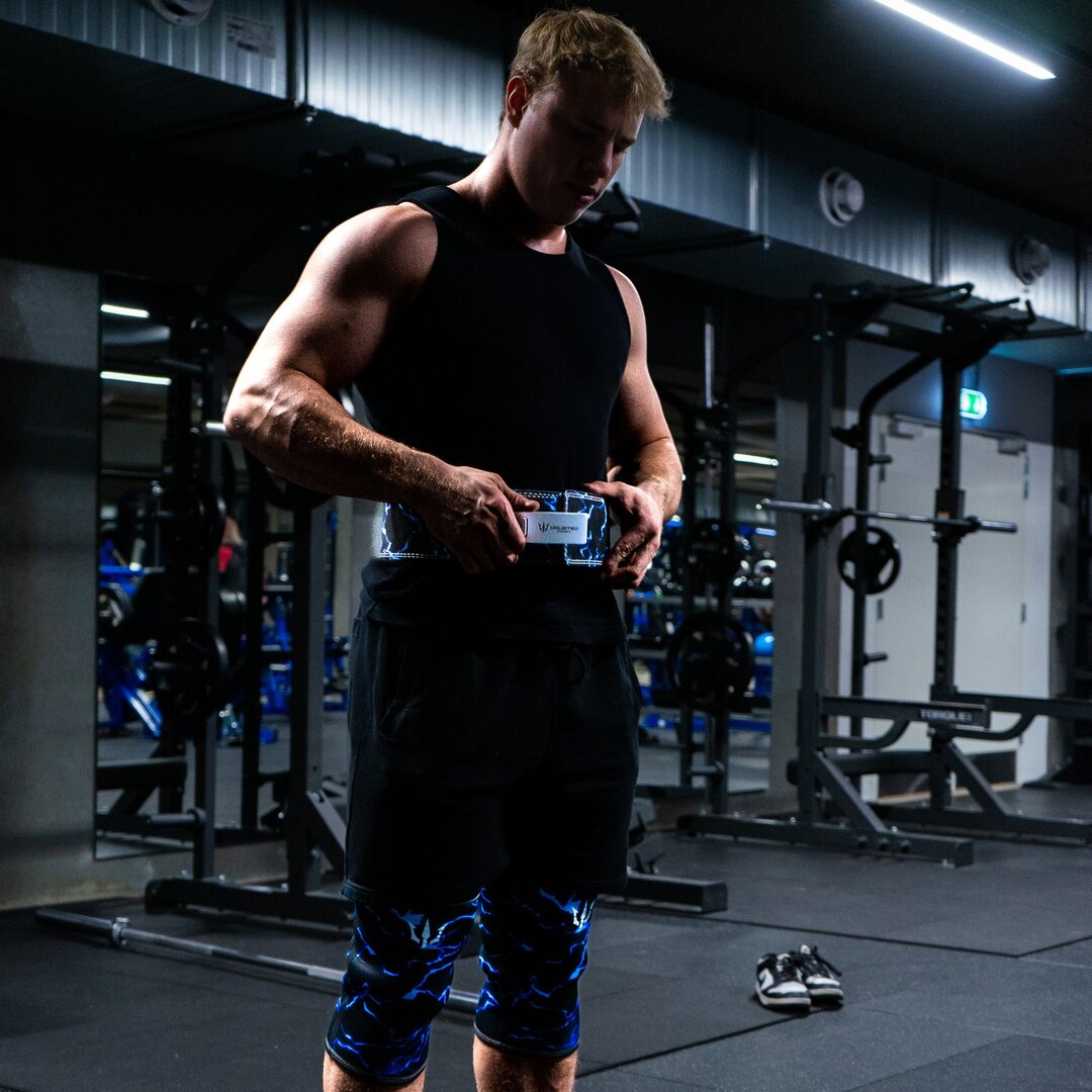 Mand spænder blå Unlisted Strength træningsbælte og har blå knee sleeves på i et fitnesscenter
